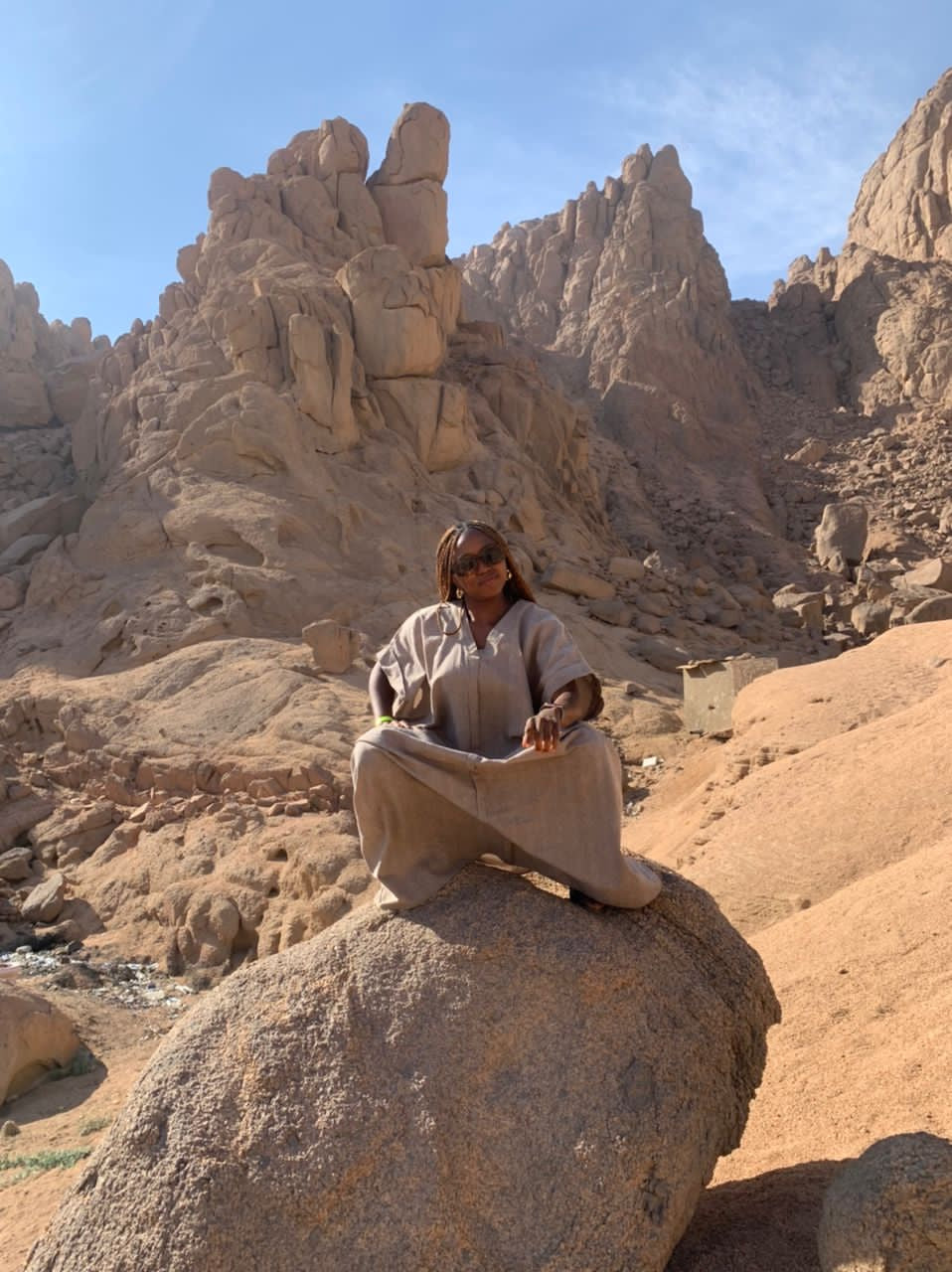 Person in loose beige clothing sitting on a rock in a sunny, rocky desert landscape