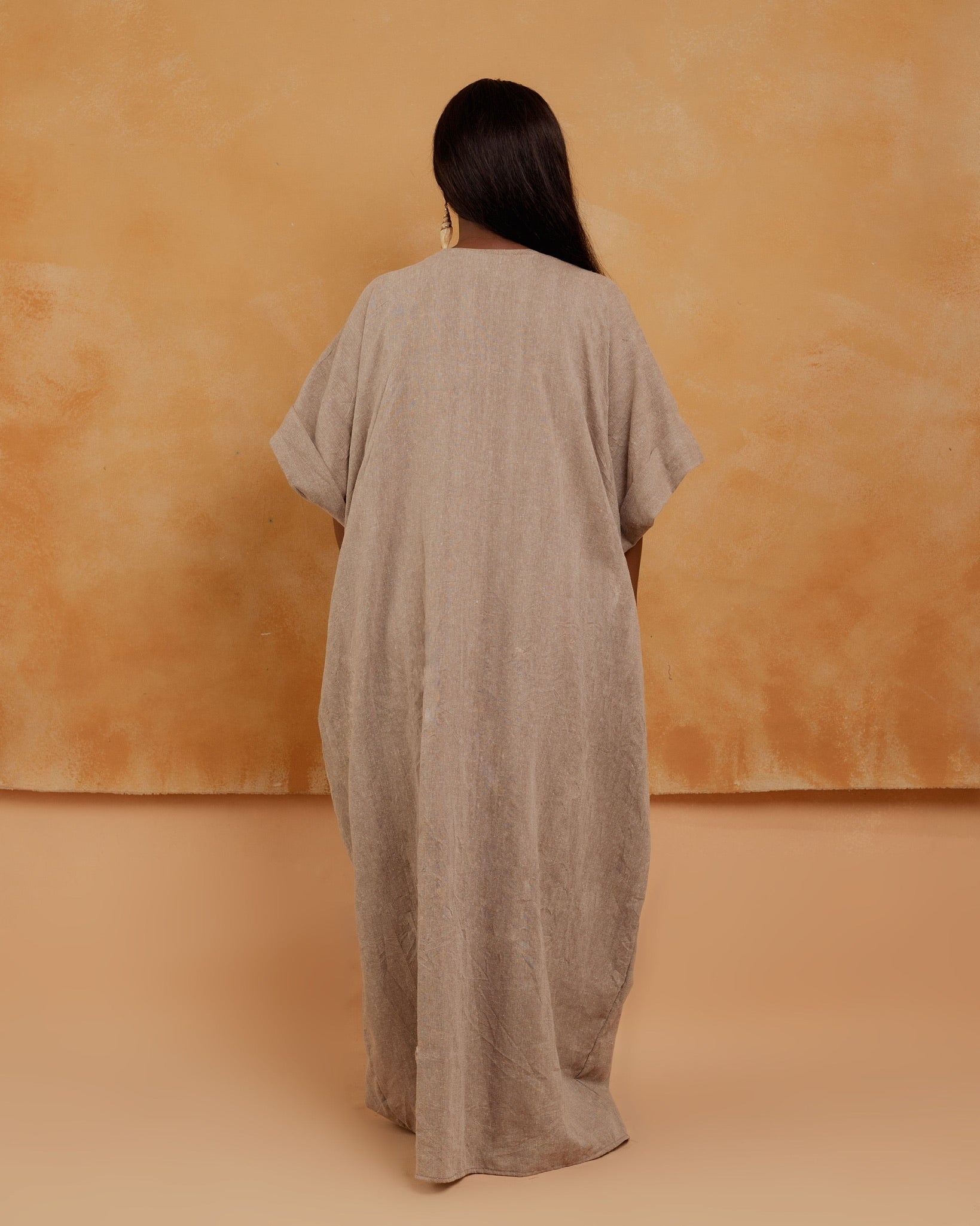 Woman in loose beige kaftan dress facing away, minimal studio background