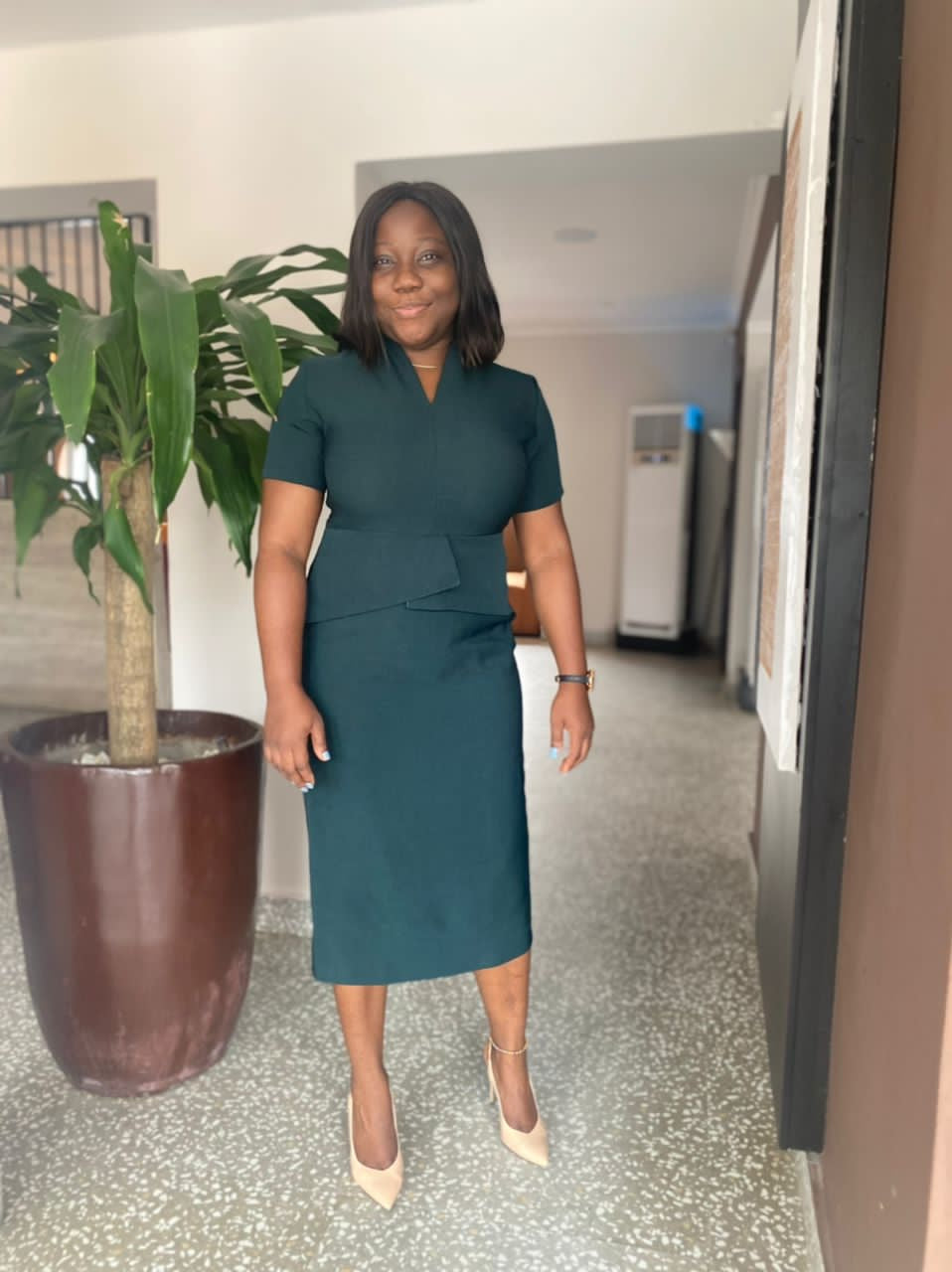 Woman in tailored green midi dress and nude heels standing indoors near a potted plant