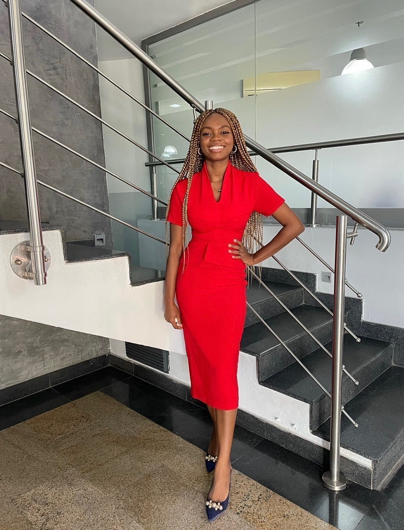 Smiling woman in a fitted red dress with braids, standing by modern staircase indoors
