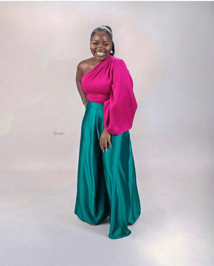 Smiling woman wears vibrant pink one-shoulder top and teal wide-leg trousers, studio backdrop