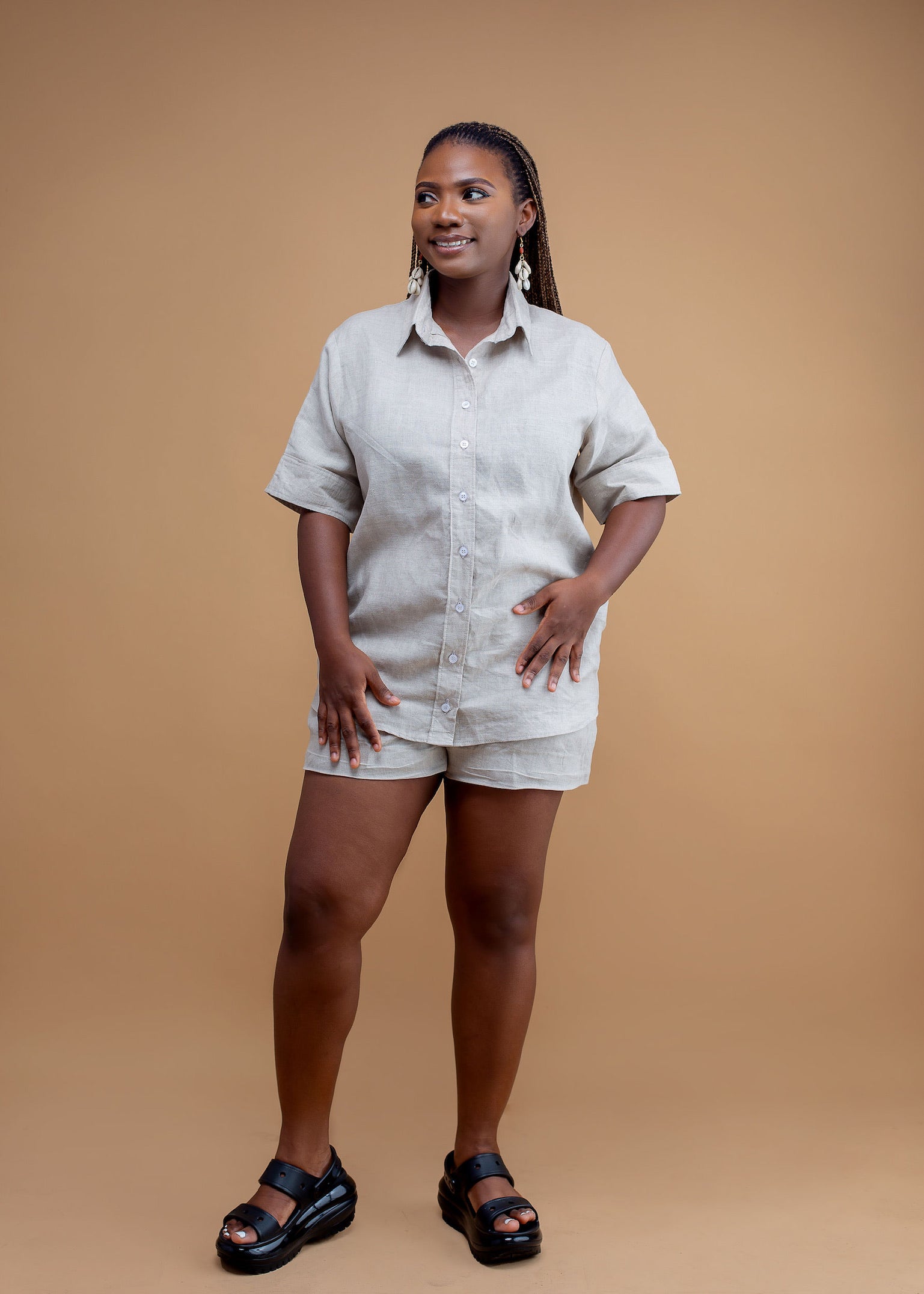 Woman wearing beige linen shirt and shorts set with black sandals on a tan background