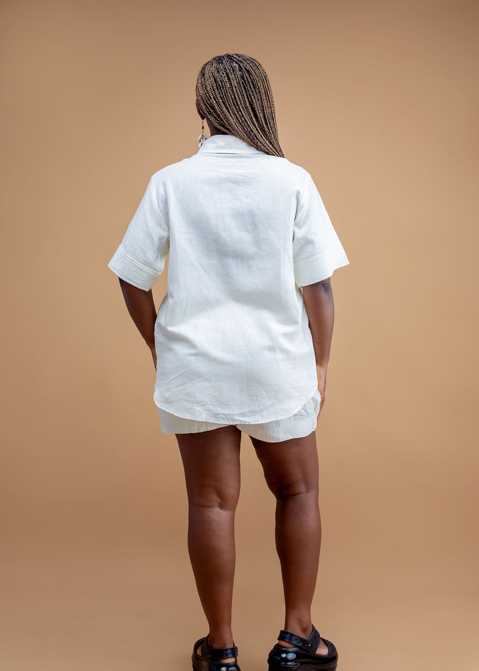Woman in white linen shirt and shorts, standing against tan background, rear view