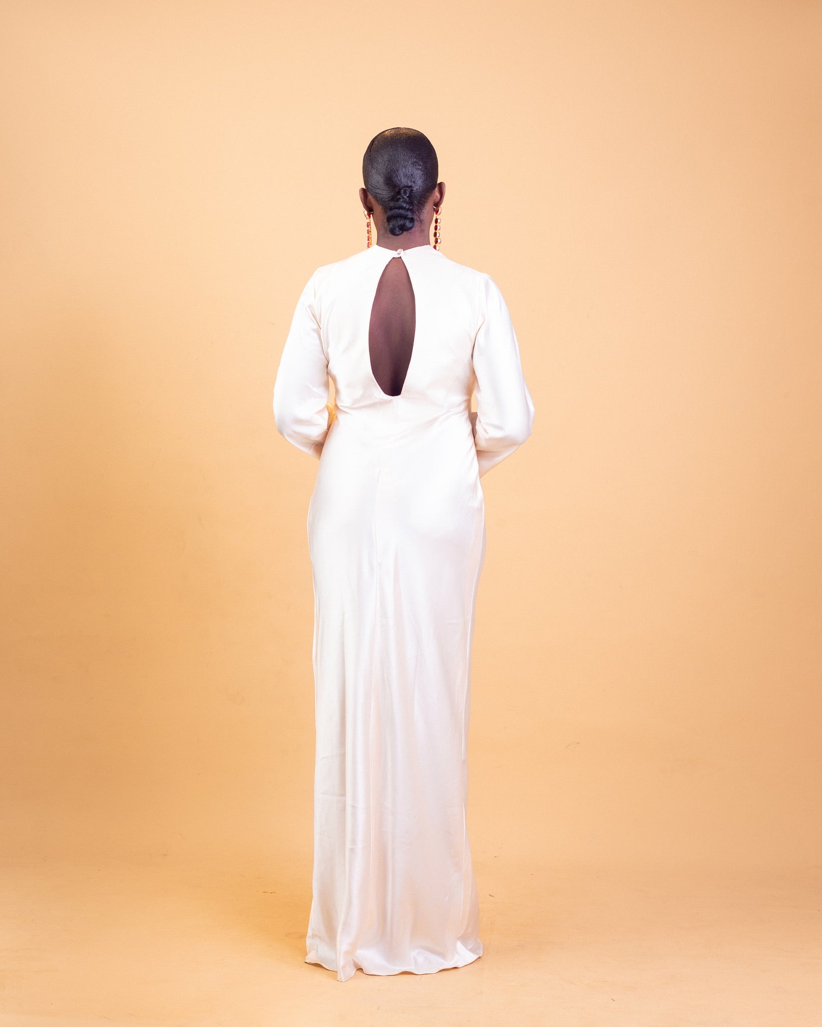 Woman in elegant white long dress with keyhole back, standing against peach background