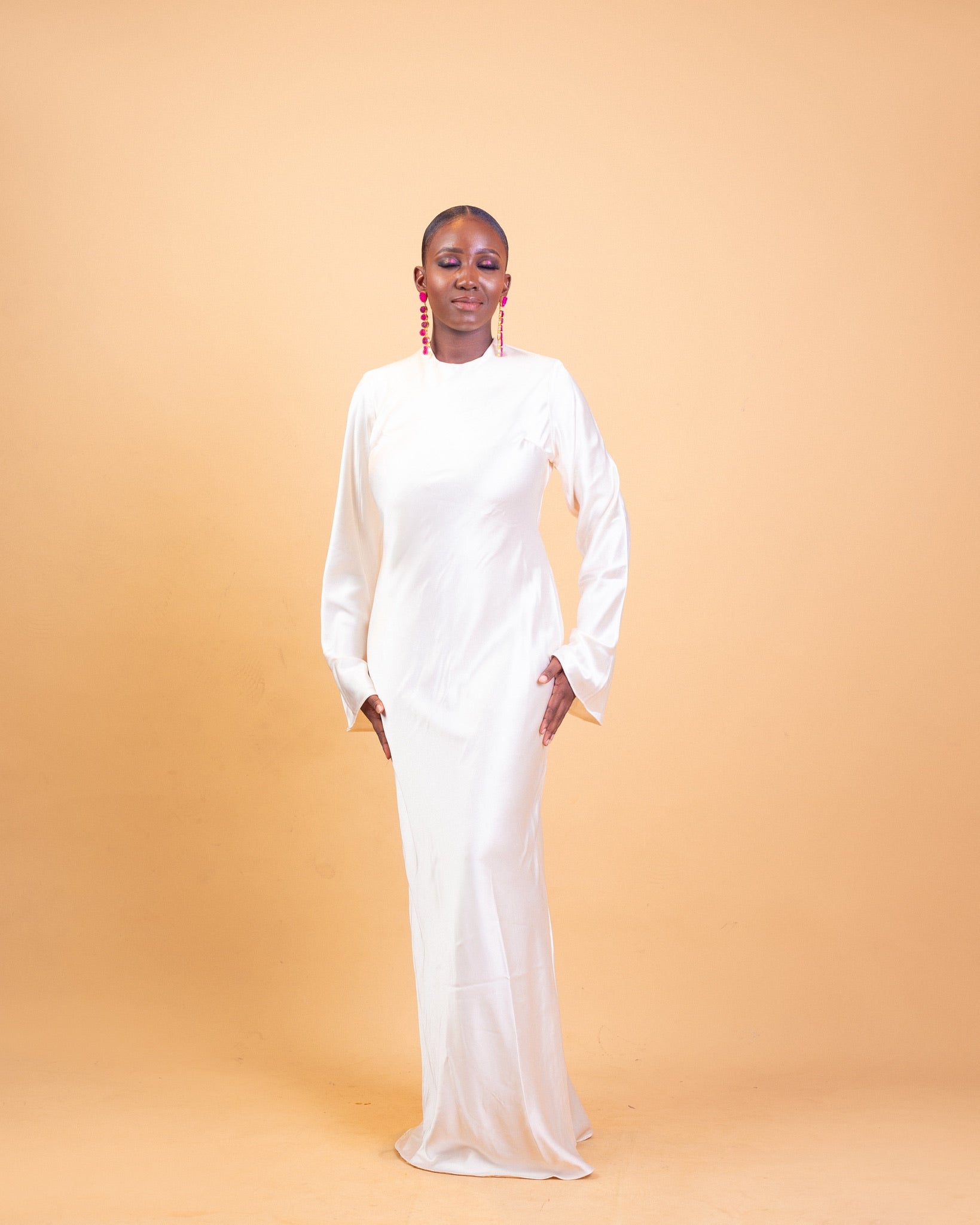 Woman in a long white fitted dress with long sleeves, posing against a peach background