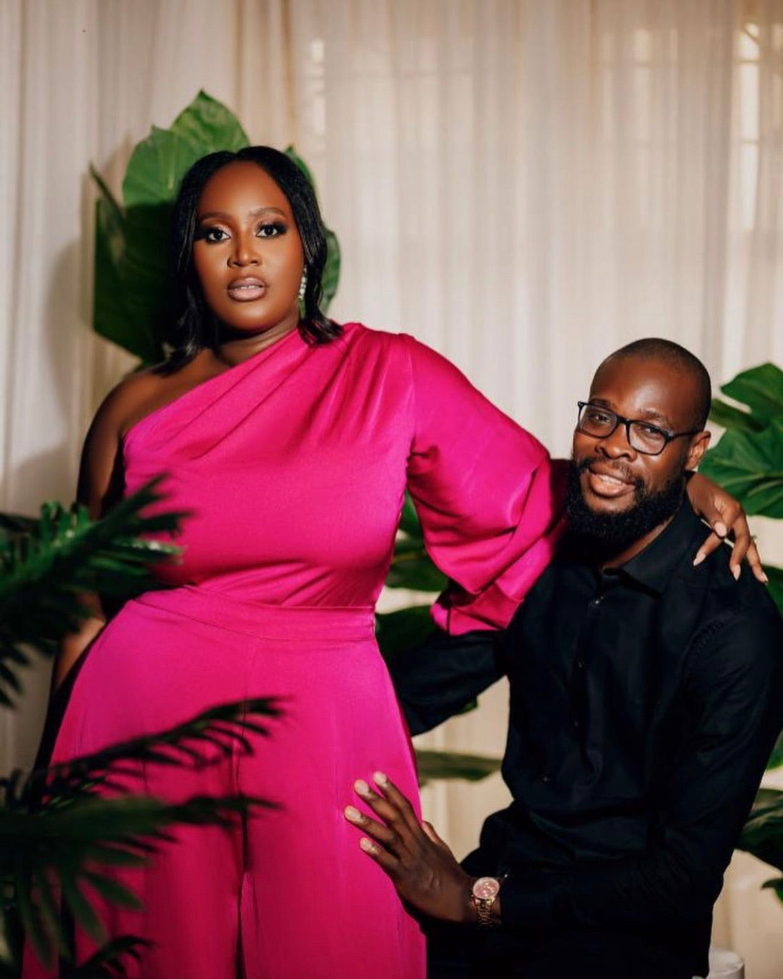 Woman in one-shoulder fuchsia jumpsuit and man in black attire posing indoors with plants