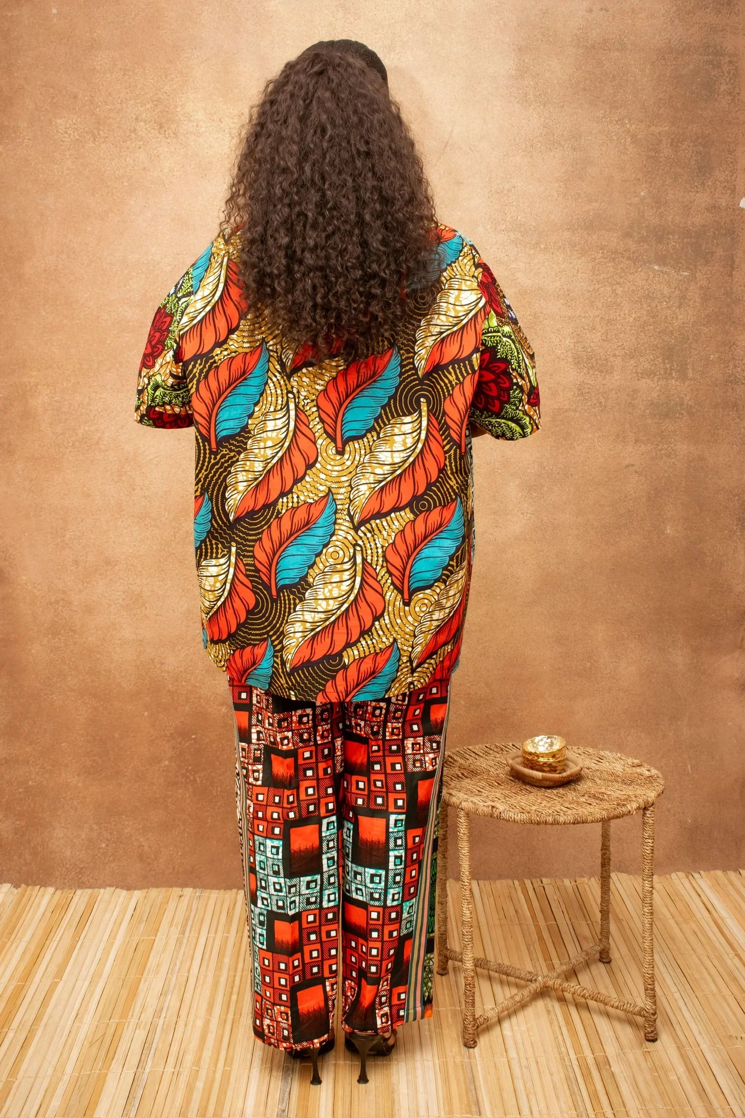 Woman in colourful Ankarza print outfit, back view, with curly hair and woven stool