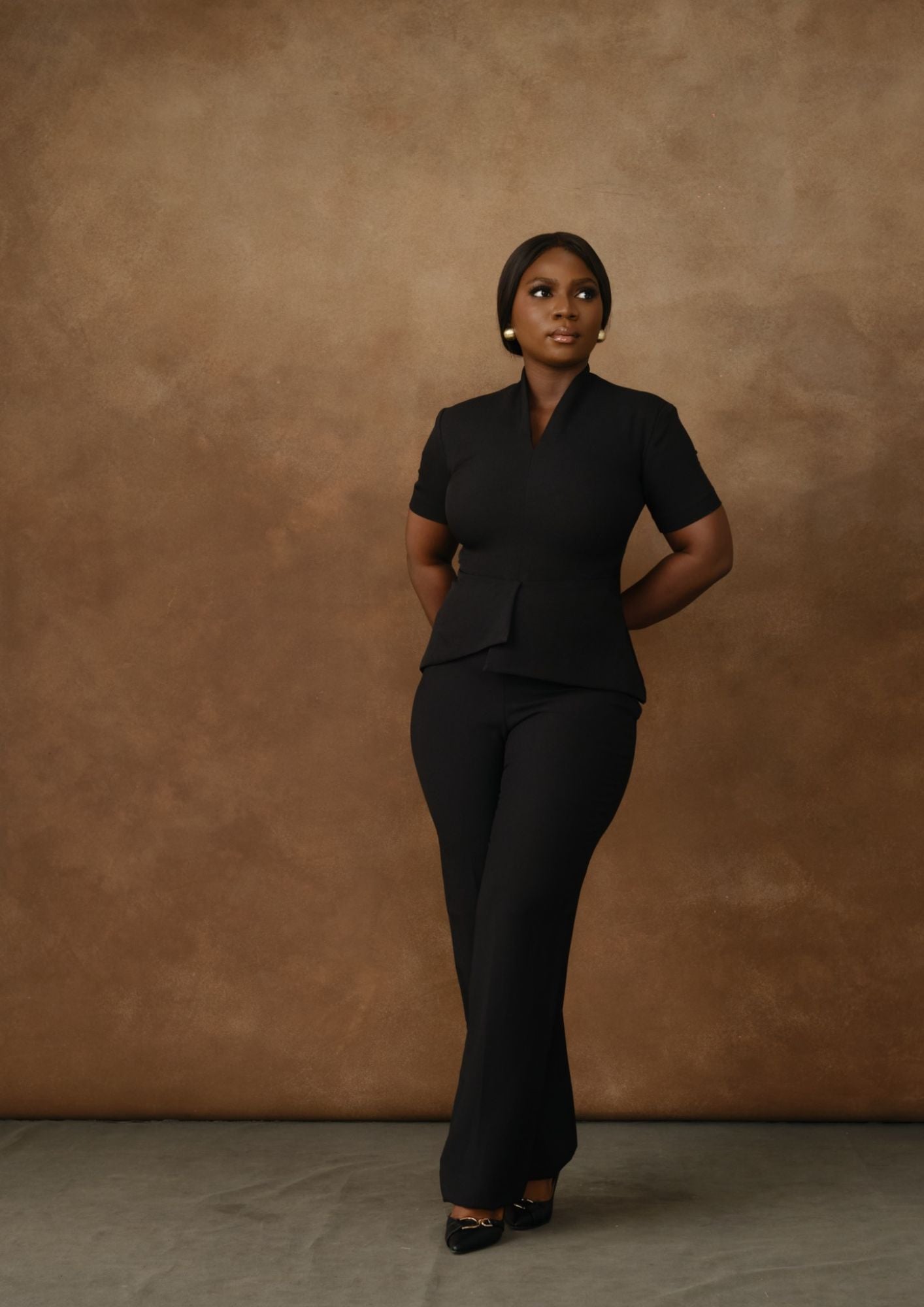 Woman wearing black tailored trouser suit, posing against a brown studio backdrop.