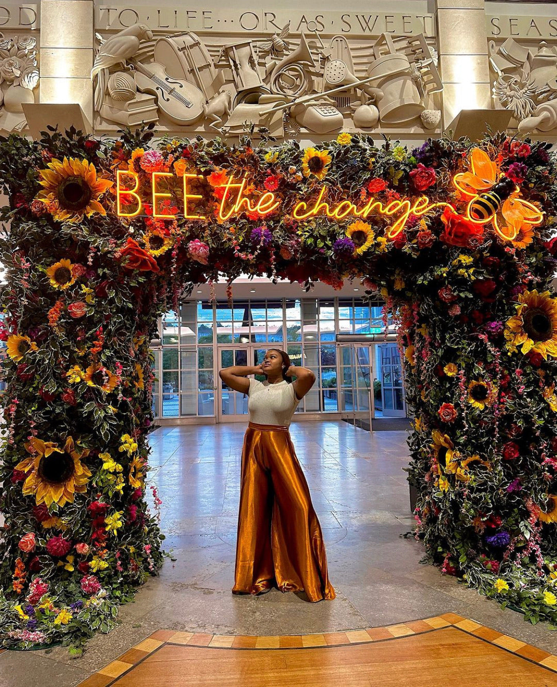 Woman in shiny orange wide-leg pants and white top posing under a floral arch with sunflowers and neon sign.