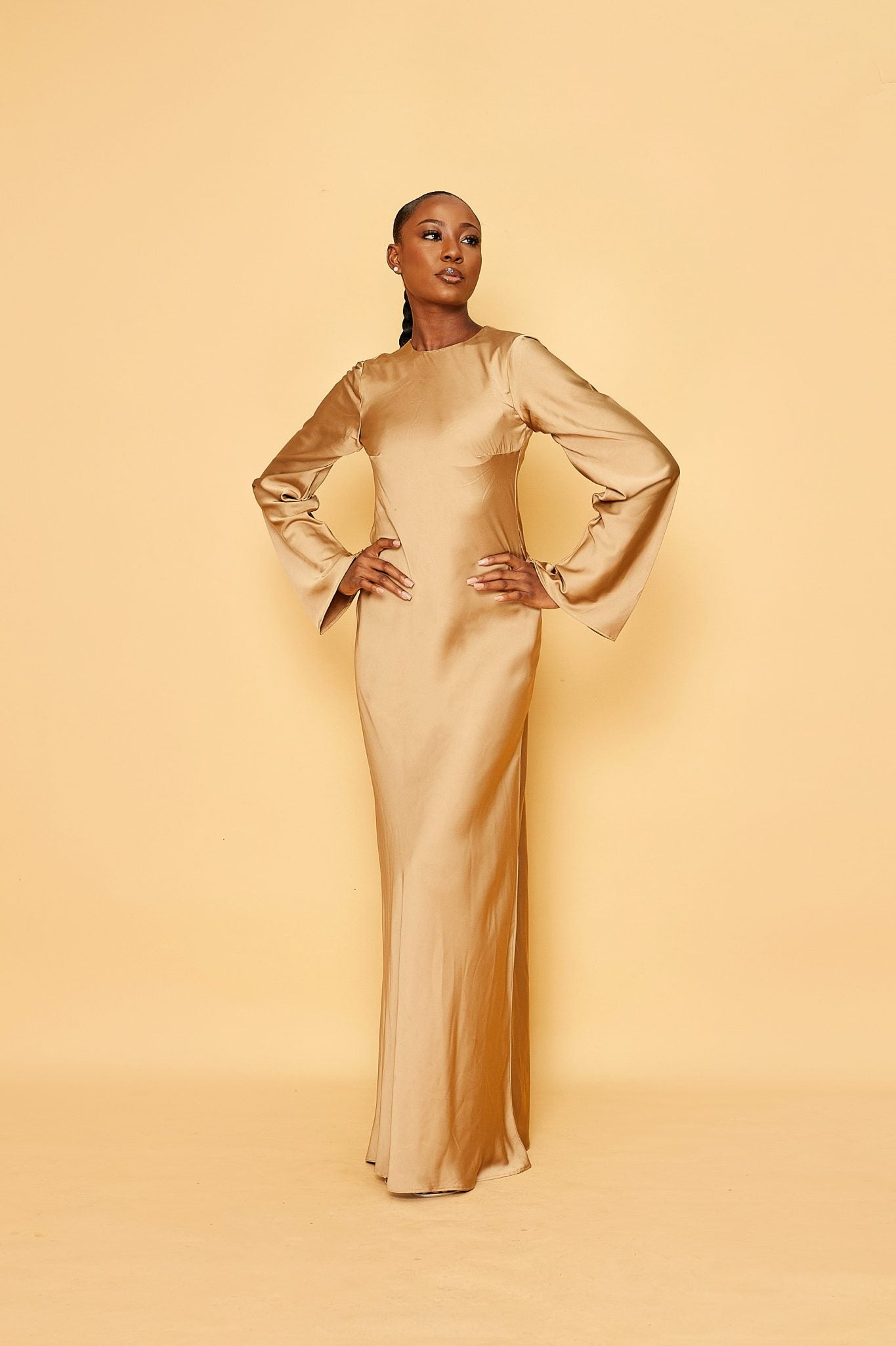 Woman in gold satin long-sleeve gown standing against a beige background, fashion portrait