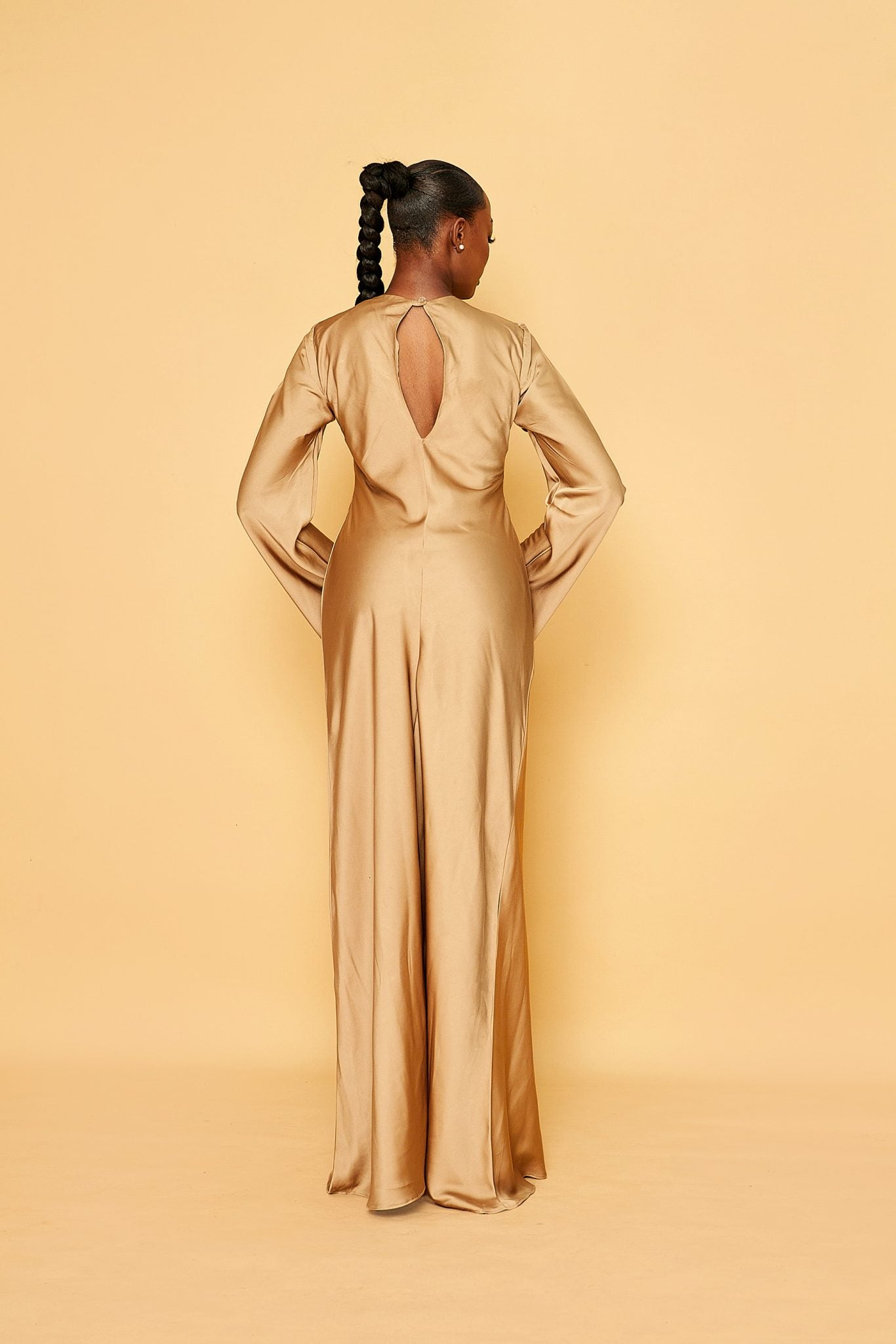 Woman in gold satin long-sleeve gown with back keyhole, braided ponytail, neutral background