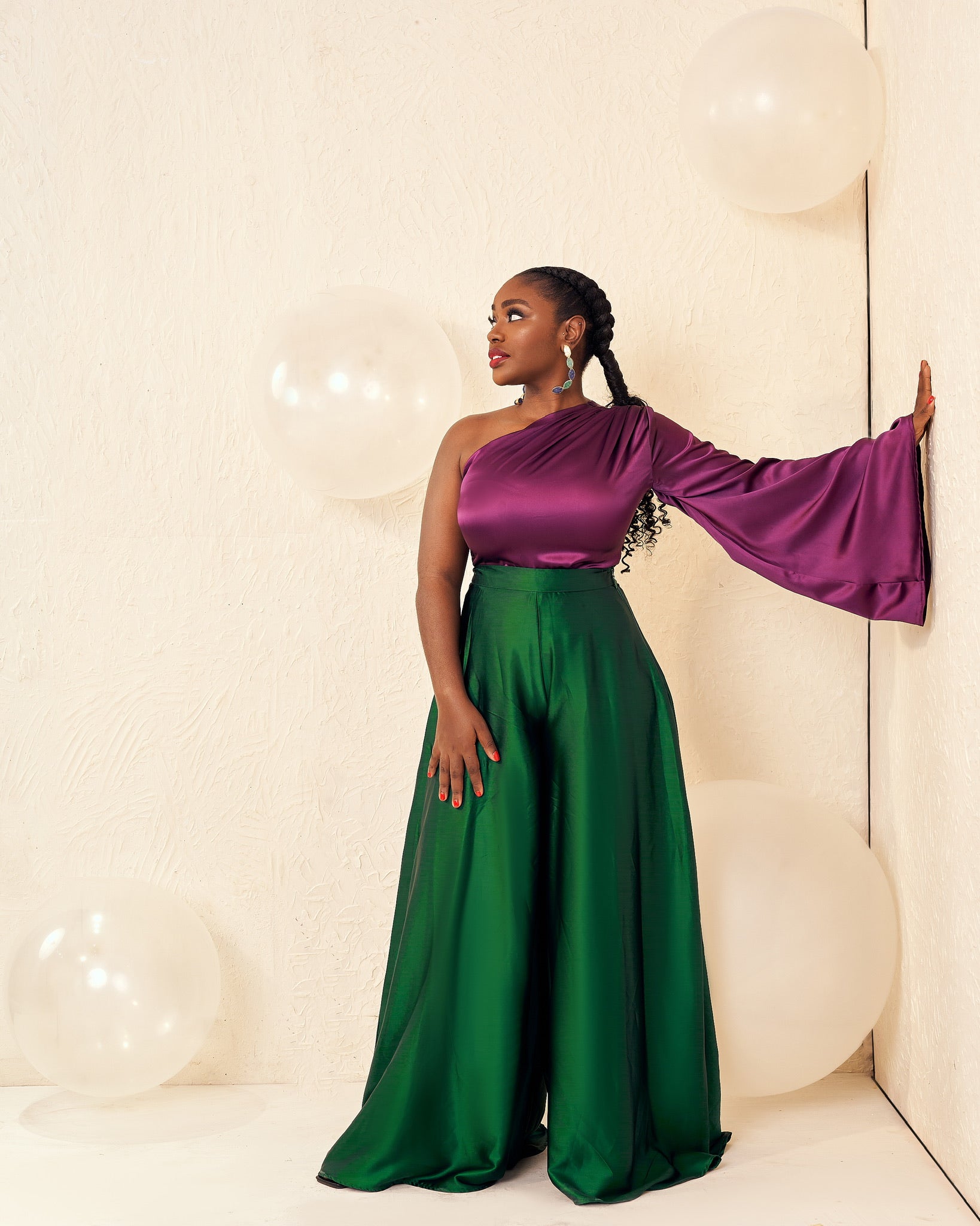 Woman in a purple one-shoulder top and green wide-leg pants, standing by a wall with white balloons.