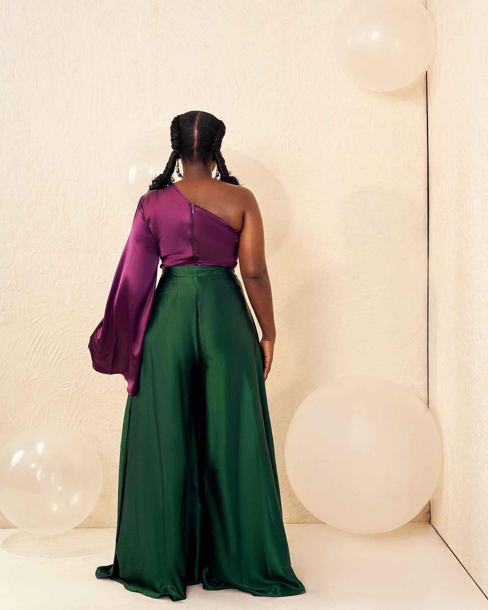 Woman in elegant purple one-shoulder top and wide-leg green pants, standing with balloons